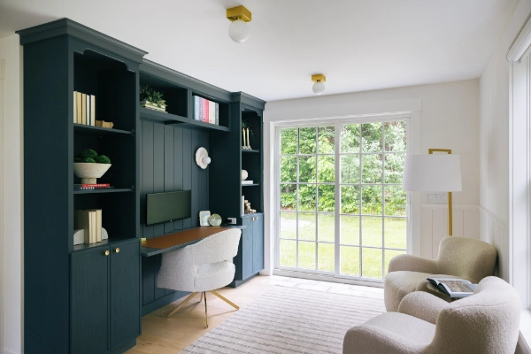 Library workspace with custom dark shelving, built-in desk, upholstered chair, and sliding glass doors opening to a green lawn